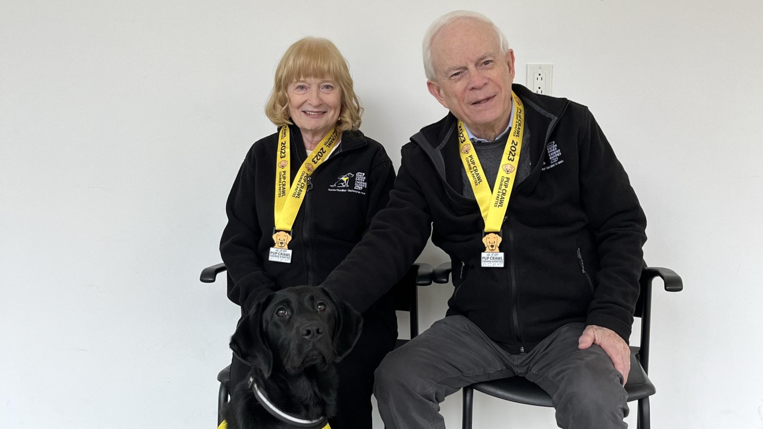 Mary and John Crocker sit to pose wearing 2023 Pup Crawl medals. They are accompanied by a black Labrador Retriever wearing a CNIB Guide Dogs vest.